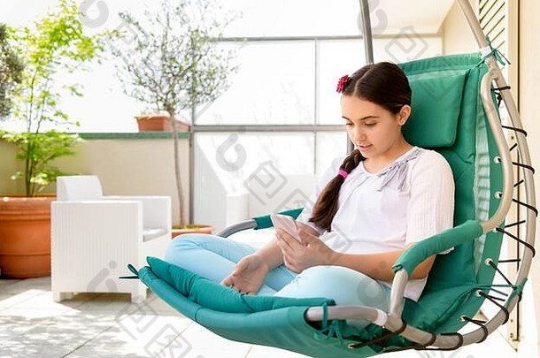 女孩坐着看媒体移动电话放松挂椅子在户外阳光明媚的天井盆栽植物