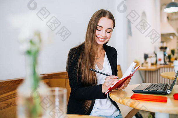 年轻的微笑女商人使笔记记事本坐着前面移动PC咖啡馆准备写信息