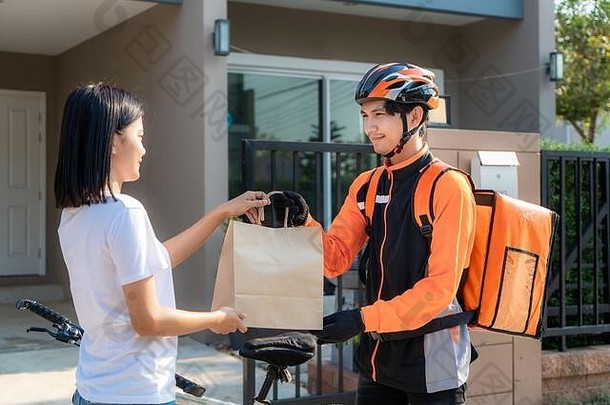亚洲男人。快递自行车交付食物橙色统一的微笑持有食物袋前面房子亚洲女人接受交付盒子