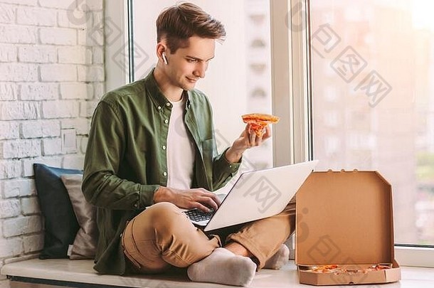 年轻的赶时髦的人男人。自由职业者吃片美味的披萨坐窗台上移动PC研究距离学习商人吃外卖