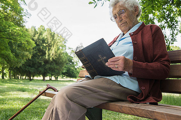 高级女人坐着公园板凳上阅读圣经
