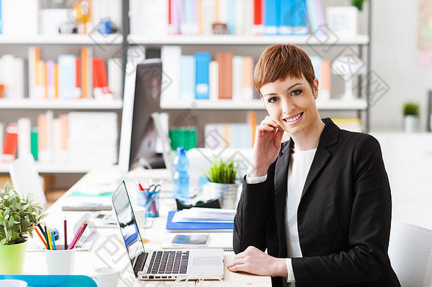自信成功的女商人坐着办公室桌子上工作移动PC微笑相机