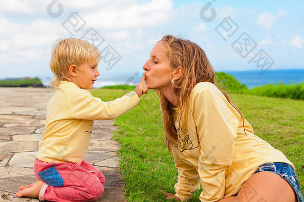 快乐家庭走孩子坐着绿色草草坪上有趣的年轻的有吸引力的妈妈。吻<strong>儿子</strong>手指积极的人活动假期