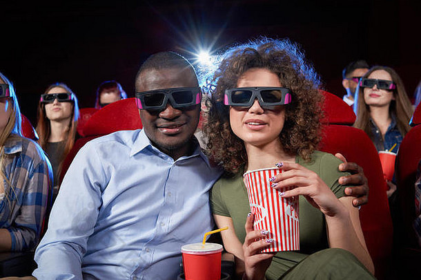 夫妇非洲式发型男人。浅黑肤色的女人女人坐着电影吃爆米花看喜剧英俊的男人。拥抱美丽的女朋友坐着电影审查