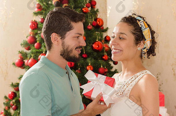 的男朋友将圣诞节现在女人女朋友快乐微笑圣诞节树装饰背景红色的礼物盒子