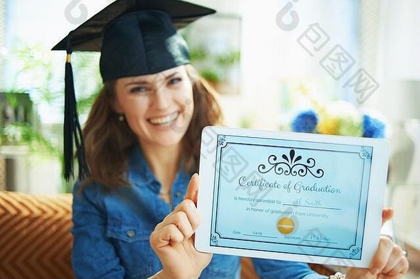 特写镜头快乐女人牛仔裤衬衫毕业帽显示电子证书毕业平板电脑屏幕生活房间阳光明媚的