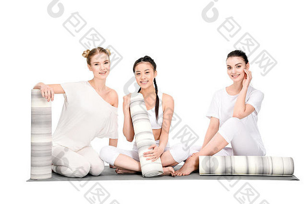 快乐多民族女孩休息瑜伽培训坐着瑜伽垫相机孤立的白色