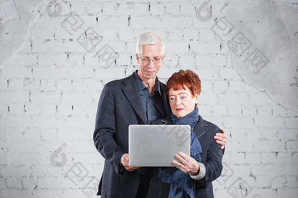人持有移动PC沟通互联网快乐微笑爷爷奶奶夫妇站拥抱孤立的白色砖背景