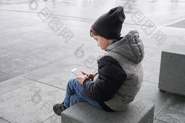 坦诚的照片年轻的男孩智能手机移动细胞电话坐着城市街冬天