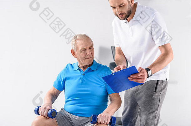 康复治疗师记事本协助高级男人。锻炼哑铃灰色背景
