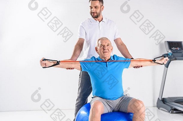 康复治疗师协助高级男人。锻炼橡胶磁带健身球灰色背景