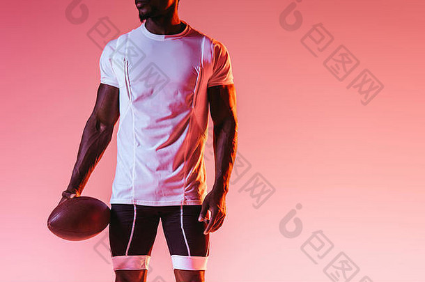 裁剪视图非洲美国运动员持有橄榄球球粉红色的背景梯度照明