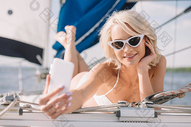 微笑有吸引力的女人比基尼太阳镜采取自拍智能手机铺设游艇