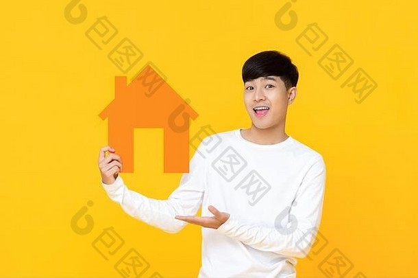 微笑英俊的亚洲男人。持有房子标志手展示手势孤立的黄色的背景
