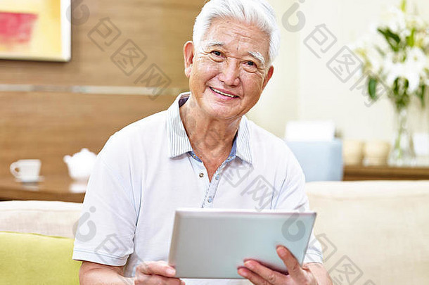 高级亚洲男人。坐着沙发持有平板电脑电脑相机微笑