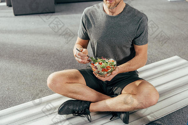 裁剪拍摄运动男人。运动服装坐着瑜伽席持有碗蔬菜沙拉首页