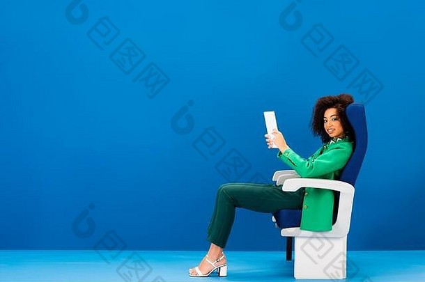 微笑非洲美国女人坐着座位持有数字平板电脑蓝色的背景