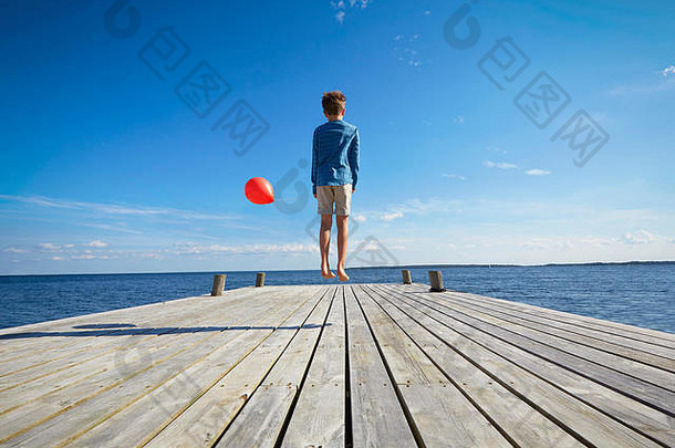 年轻的男孩跳木码头持有红色的氦气球后视图