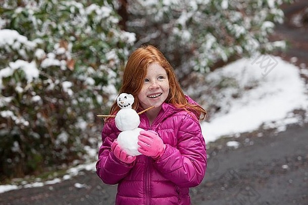 红色的头发的女孩前面雪覆盖树穿粉红色的手套垫夹克持有雪人微笑