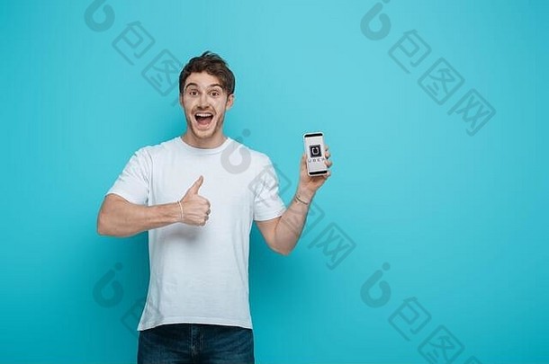 基辅乌克兰4月兴奋年轻的男人。显示拇指持有智能手机超级应用程序屏幕蓝色的背景