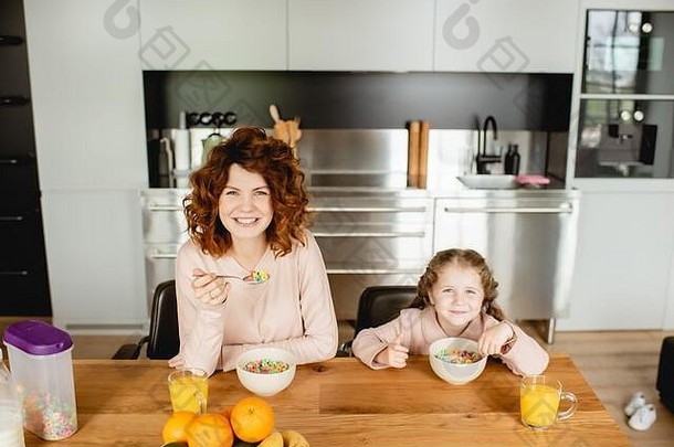 快乐妈妈。可爱的女儿持有勺子碗玉米片眼镜橙色汁