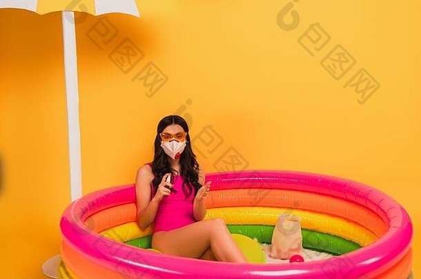 年轻的女人泳衣太阳镜医疗面具坐着充气池持有洗手液黄色的