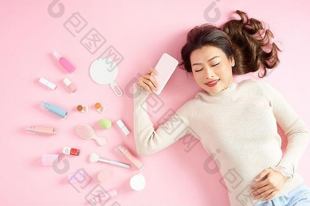 亚洲女人持有电话说谎粉红色的背景化妆品工具