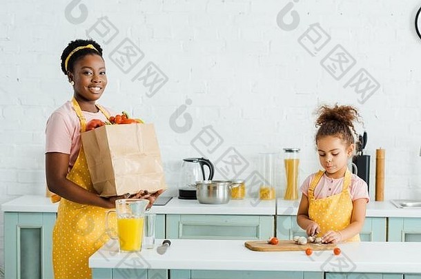 快乐非洲美国妈妈。持有纸袋食品杂货女儿切割蘑菇