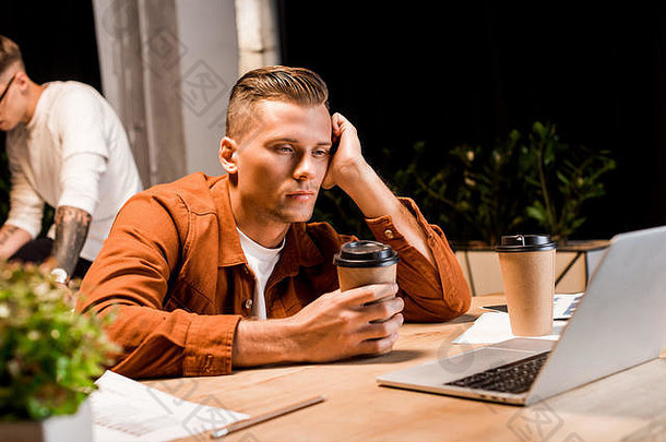 年轻的疲惫商人持有咖啡坐着移动PC晚上办公室