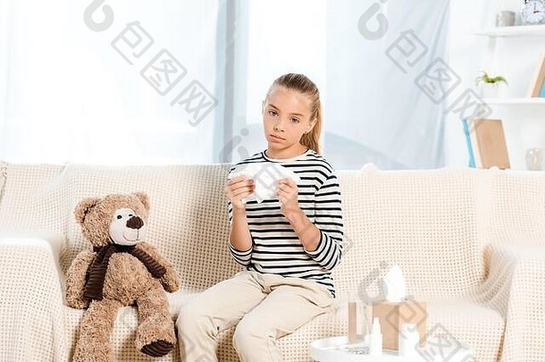 生病的孩子持有餐巾泰迪熊坐着沙发