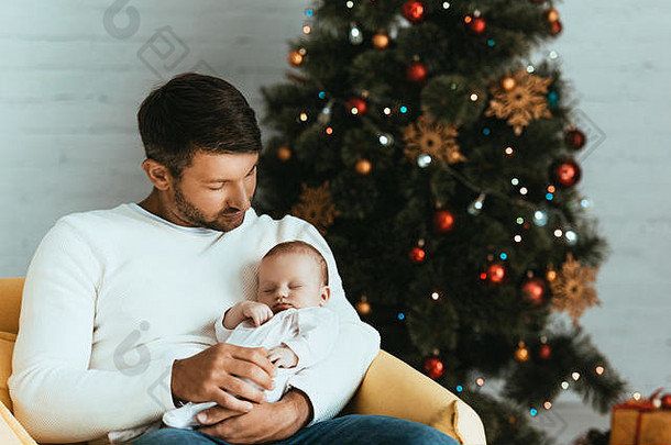 快乐父亲持有睡觉婴儿坐着圣诞节树