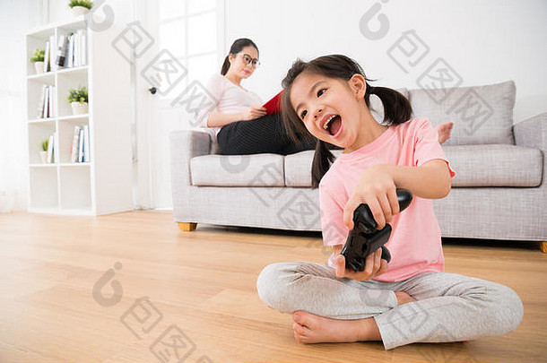 年轻的妈妈。沙发研究女儿快乐持有操纵杆玩视频游戏木地板上享受放松家庭假期