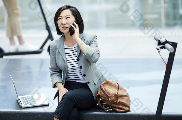 cold-toned肖像现代亚洲女人说话智能手机坐着混凝土板凳上在户外电踏板车前景复制空间