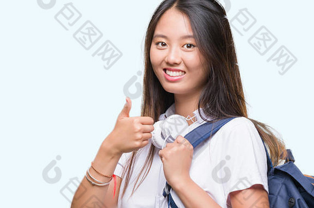 年轻的亚洲女人穿背包耳机孤立的背景快乐大微笑标志拇指<strong>手指</strong>优秀的标志