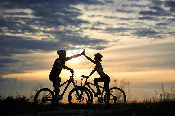 可爱的夫妇头盔采取家庭骑自行车站摆姿势轮廓运动男人。女人高令人惊异的日落美丽的天空背景