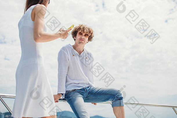 肖像高加索人英俊的卷曲的男人。坐着帆船栏杆穿太阳镜相机蓝色的天空背景Copyspace