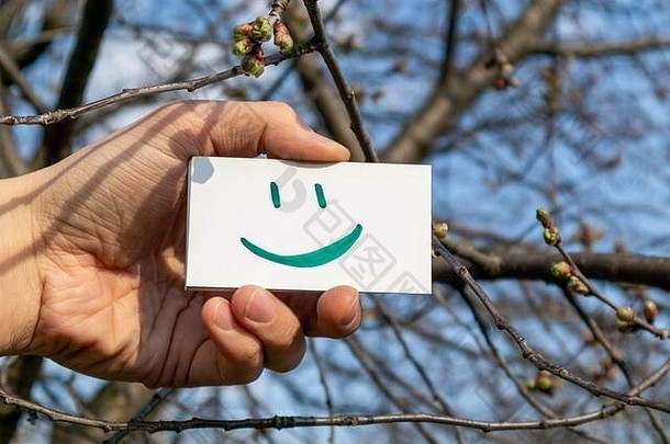 快乐绿色微笑表情符号白色纸卡持有手阳光明媚的春天蓝色的天空背景积极的情绪好天气时间