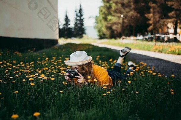年轻的美丽的女孩橙色t恤持有复古的相机手说谎草坪上蒲公英成长开花蒲公英