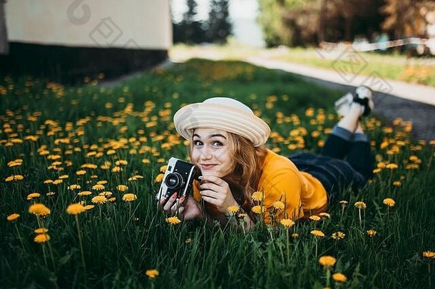 年轻的美丽的女孩橙色t恤持有复古的相机手说谎草坪上蒲公英成长开花蒲公英
