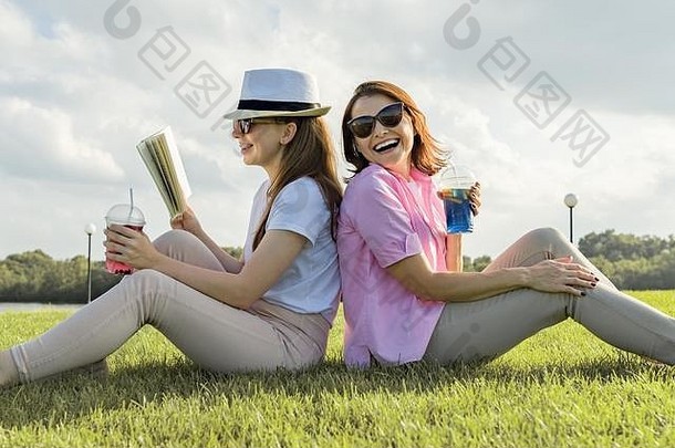 沟通父少年妈妈。青少年女儿坐着绿色草坪上公园阅读书会说话的喝很酷的鸡尾酒