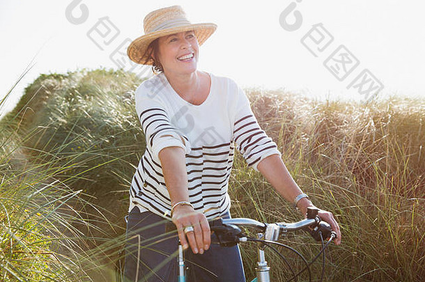 微笑成熟的女人骑自行车阳光明媚的海滩草路径