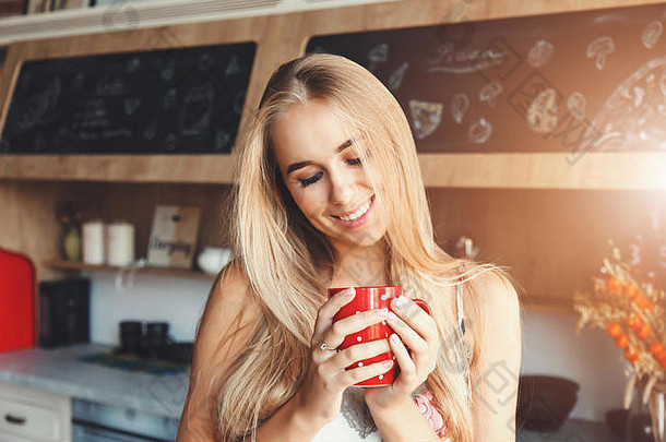 不错的金发女郎长头发女孩穿斯捷装站红色的阁楼舒适的阳光明媚的厨房气候变暖一个人的手杯咖啡欢乐一天
