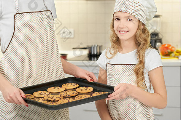 前面视图微笑女孩烹饪他围裙相机摆姿势妈妈。保持托盘饼干厨房孩子烘焙美味的纸杯蛋糕帮助妈妈。