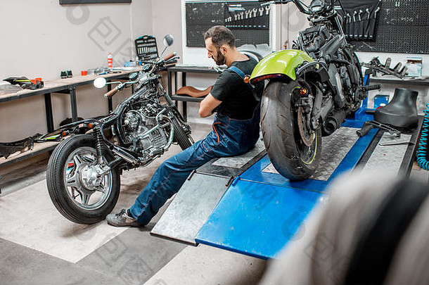 肖像英俊的有胡子的骑摩托车的人工作工作服坐着摩托车维修车间