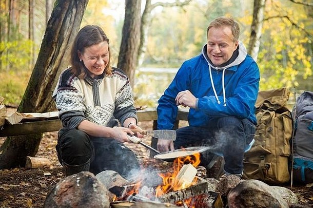 男人。女人使煎饼篝火森林海岸湖使火烧烤快乐夫妇探索芬兰斯堪的那维亚景观