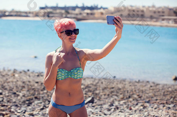旅行休闲夏天技术人概念年轻的女人粉红色的短头发采取自拍海滩背景
