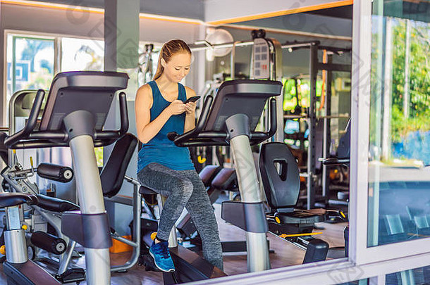 年轻的女人电话培训健身房女人坐着锻炼机持有移动电话