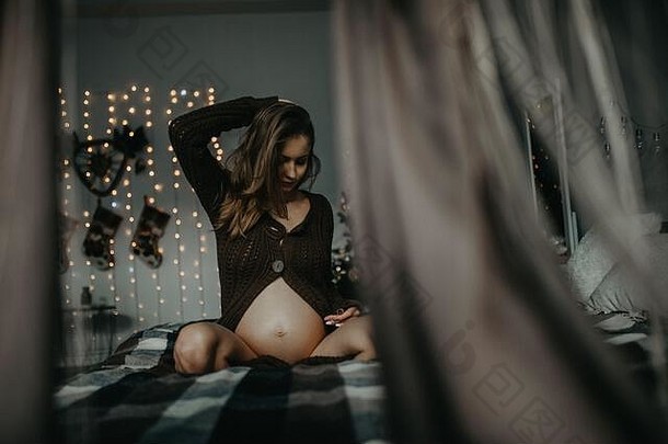 年轻的怀孕了女人坐在床上背景<strong>圣诞</strong>节树装饰视图窗帘