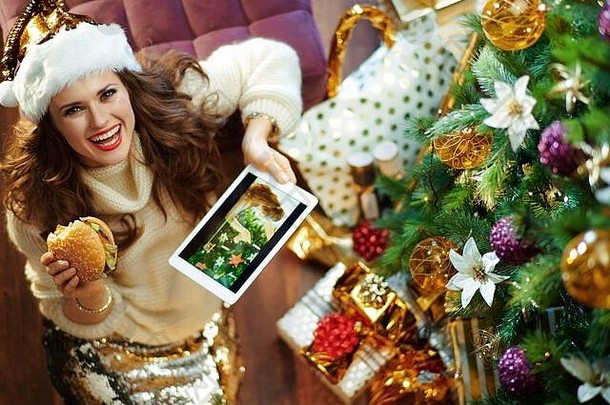 微笑现代女人长浅黑肤色的女人头发黄金金片裙子白色毛衣装饰圣诞节树现在盒子吃汉堡