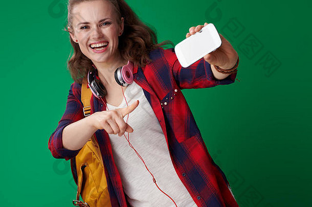 微笑年轻的学生女人红色的衬衫背包耳机指出智能手机绿色背景